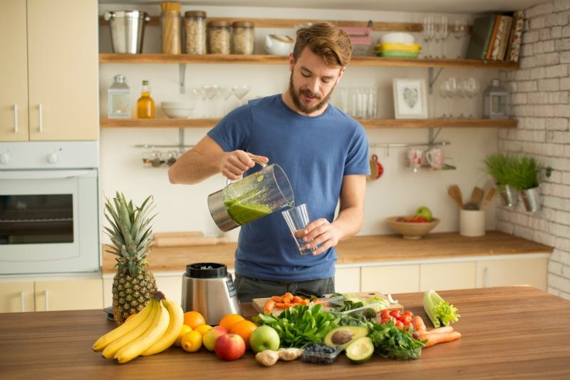 A bearded man in a kitchen pours a green smoothie into a glass, surrounded by fresh fruits and vegetables on a countertop.