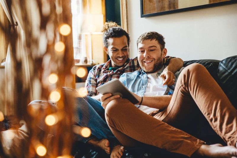 A happy couple sits on a couch, one holding a tablet as they laugh together in a cozy room, softly illuminated by string lights in the foreground.