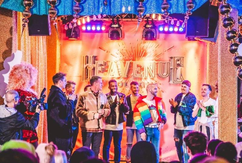 A group of people celebrating on a glittering stage with a golden 'Heavenue' sign in the background, surrounded by festive decorations.