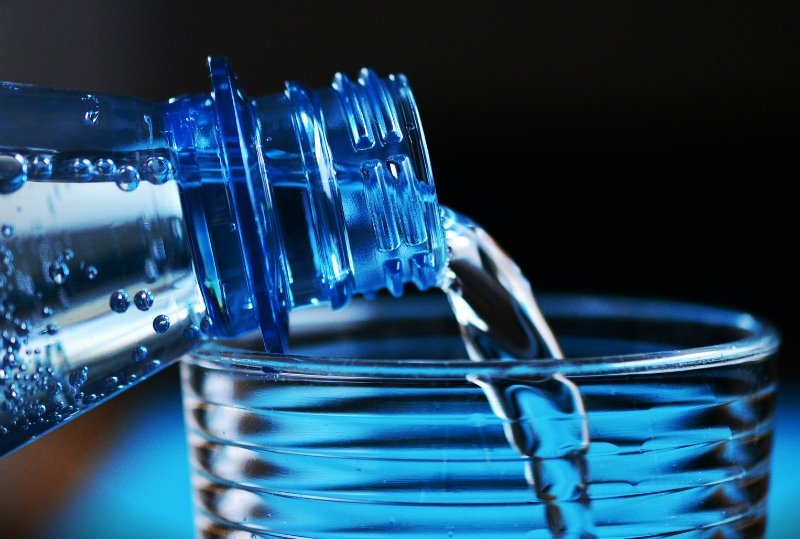 A close-up of water being poured from a blue bottle into a glass, with droplets visible on the rim and a refreshing, clear liquid.